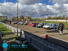 Ряд барвистих гоночних автомобілів і водіїв на сонячній гоночній трасі, на тлі яскравих осінніх дерев і розсіяних хмар. Атмосфера жвава і конкурентна.