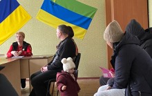 Представниця Лиманської МВА сидить за столом та розповідає присутнім ВПО громади про послуги у новому хабі.