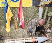На полу лежить уламок снаряду, хлопець уважно розглядає. Позаду нього прапори бригад та захисна сітка зеленого кольору та тубуси.