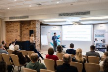 Учасник форуму,стоїть біла екрану та розповідає про презентацію. Іньші учасники слухають.