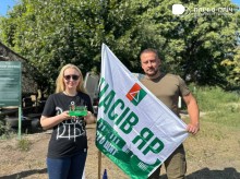 Чоловік та жінка стоять на вулиці, позаду зелені дерева. Чоловік тримає прапор Часовоярської громади.