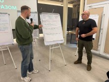 Обговорення робочих процесів.  Молодий чоловік з мікрофоном щось розповідає та показує рукою на мольберт, поруч стоїть начальник міської військової адміністрації Сергій Чаус.