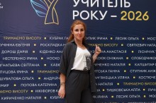 Ганна Никитська посміхається та показує жест «вгору» на тлі темно-синього банера конкурсу «Учитель року – 2026» з іменами учасників.
