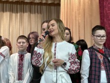 Дія на сцені. Дівчина у національному вбранні тримає мікрофон, позаду стоять хлопці та дівчата теж у національному вбранні.