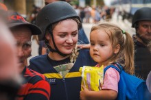 Жінка-рятівник у шоломі ніжно тримає на руках маленьку дівчинку, яка тримає жовту пакунку. Ця сцена передає тепло і безпеку на тлі багатолюдного фону.