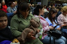 Учасники заходу сидять у залі. Жінка тримає немовля.