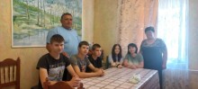 Великий стіл за столом сидять хлопці, дівчата та жінка. Чоловік стоїть біля стіни