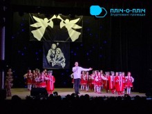 Дія на сцені. Актор тримає у руках мікрофон та співає, розводить рукою. З обох боків стоять діти у народних костюмах червоного кольору.