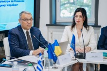 Крупний план двох учасників конференції: чоловік в окулярах та синьому піджаку виступає перед мікрофоном, поруч з ним сидить жінка у білому жакеті.
