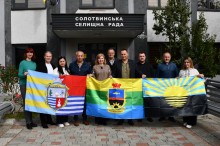 Група з десяти осіб стоїть перед будівлею з вивіскою українською мовою. Вони тримають три кольорові прапори з емблемами, посміхаються і дивляться в камеру. Атмосфера офіційна, але дружня.