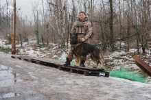 Дія на вулиці. На дерев'яних піддонах стоїть військовий та поруч пес Арчі.
