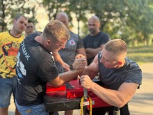 Двоє чоловіків змагаються, один схиляє руку противника. Троє чоловіків спостерігають за турніром.
