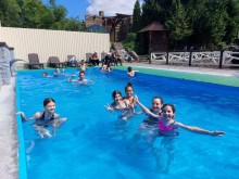 Дівчата та хлопці у басейні, посміхаються та махають руками. Вода у басейні блакитного кольору, біля басейну стоять шезлонги.