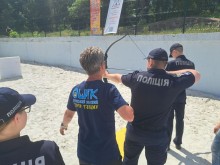 Інструктор Донецького обласного центру туризму навчає курсанта у формі поліції основам стрільби з лука на спортивному майданчику.
