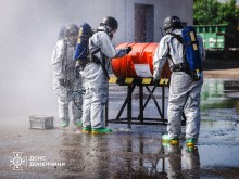 Рятувальники у захисних костюмах на вулиці. Стоять біля помаранчевого контейнера. Навчаються ліквідовувати наслідки витіку хімічної зброї.