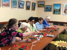Жінки сидять за столом, на якому розкладені стрічки, ножиці та клейові пістолети, і зосереджено виготовляють квіти маку
