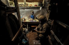 Захисник України находиться у бліндажі. На столі стоїть гріється чайник та лежіть їжа. Солдат сидить з телефоном в руках.