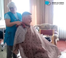 Жінка - перукар стоїть біля чоловіка, який сидить у перукарському кріслі. Жінка у синій уніформі.