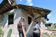 Журналіст спілкується з господинею пошкодженого будинку. На задньому плані будинок без даху та вікон.