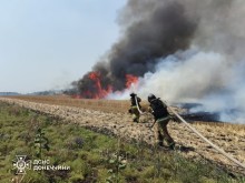 Двоє рятувальників з гідрошлангом, рухаються по полю, попереду вогонь та чорний дим.