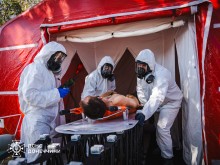 Рятувальники у спеціальних білих костюмах. Відпрацьовують техніку та допомогу постраждалим унаслідок хімічної атаки. На медичній кушетці лежить умовно потерпілий.