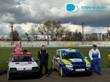 Два ралійні автомобілі припарковані на гоночній трасі. Між ними стоїть чоловік у чорному, а інший у синьому костюмі спирається на автомобіль. На тлі похмуре небо та осінні дерева.