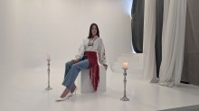 Дівчина у вишиванці, джинсах та білих туфлях сидить на кубі у фотостудії між двома свічниками