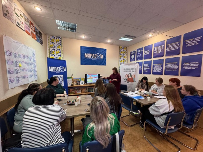 Зустріч жінок. Жінки сидять за столами, одна з учасниць стоїть біля екрану монітора та розповідає  про збереження культурної ідентичності та підтримку переселених громад та способи зберегти зв’язок із рідним краєм, проживаючи в інших містах.