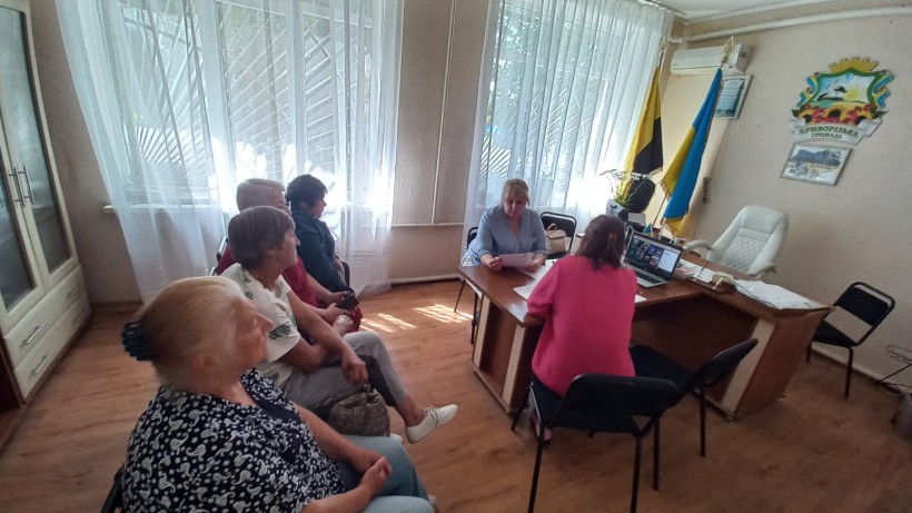 Кілька людей сидять обличчям до жінки за столом, яка, проводить онлайн-зустрічі з ветеранами та членами родин загиблих захисників і захисниць України. На столі видно ноутбук, що відображає відеодзвінок, а на стіні за столом висить український прапор.