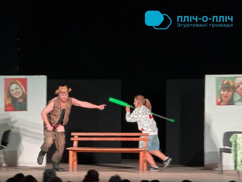 Театральна сцена: актор тікає, акторка з зеленою палицею рухається за ним; на тлі — фото на білих панелях.