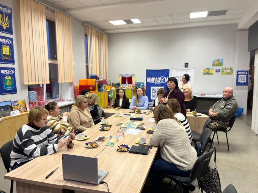 Учасники гри, сидять за столом на краю стола стоїть ноутбук,на столі солодощі,чай та гра.