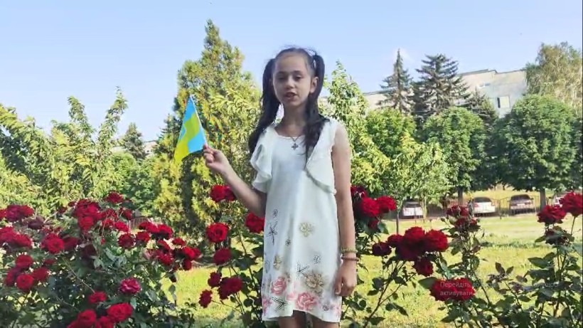 Маленька дівчинка з українським прапором на тлі квітучих троянд та зелені декламує вірш до Дня Конституції України.