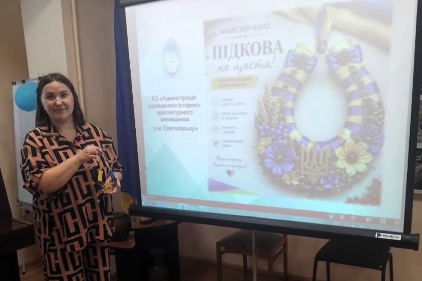 Ведуча заходу тримає декоративну підкову біля екрана з презентацією майстер-класу «Підкова на щастя!»