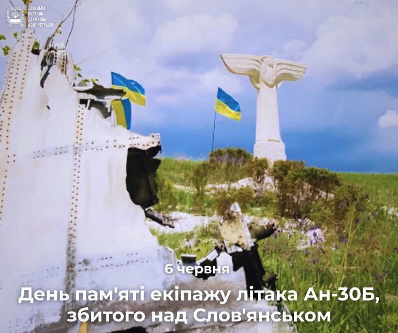 Меморіальна сцена показує уламки літака і два українські прапори на тлі блакитного неба, зі статуєю білого ангела на задньому плані, що нагадує про урочисту пам'ять.
