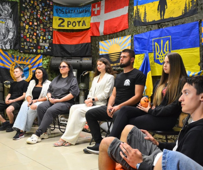 Учасники заходу сидять на стільцях, уважно слухають, позаду них - прапори різних частин ЗСУ.
