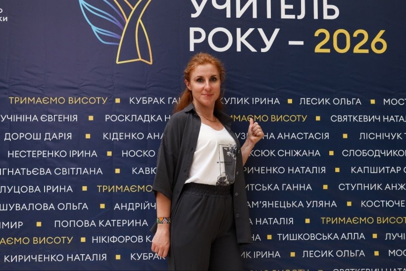 Ганна Никитська посміхається та показує жест «вгору» на тлі темно-синього банера конкурсу «Учитель року – 2026» з іменами учасників.
