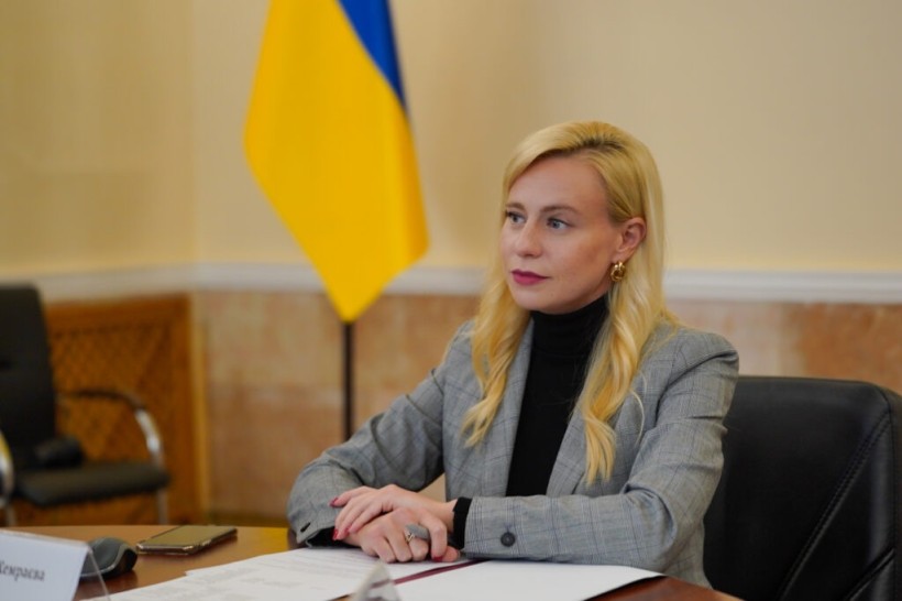 Леся Хемраєва сидить за столом на фоні прапора України