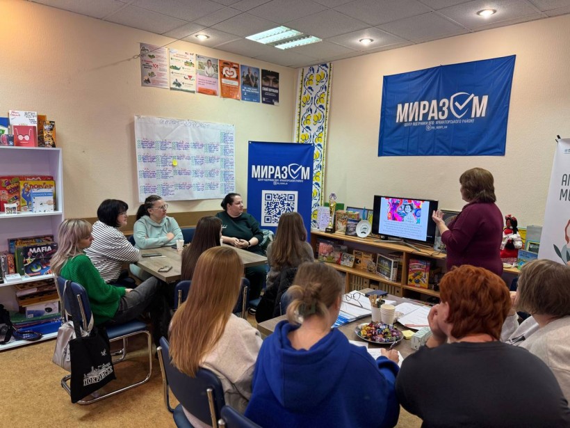 Жінки уважно слухають розповідь учасниці зустрічі, яка стоїть біля екрану монітора. На стінах висять прапори організаторів Smarta 
«МиРазом» Центр підтримки ВПО Краматорського району