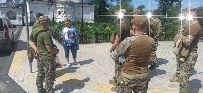 Військовослужбовці та гід на фоні стіни заповідника, пошкодженої осколками