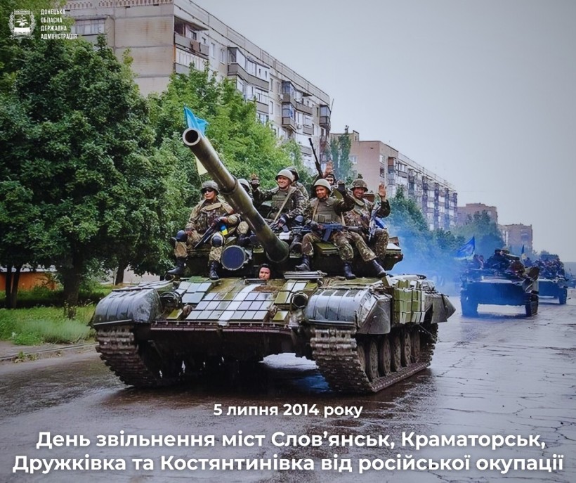 Військовий танк, що перевозить солдатів з українським прапором, їде вулицею міста, обсадженою деревами та будівлями. Текст повідомляє про річницю визволення.