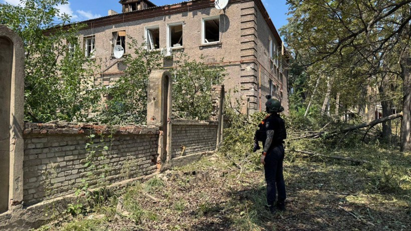 Правоохоронець стоїть біля двоповерхового будинку, в якому обстрілом вибило вікна.