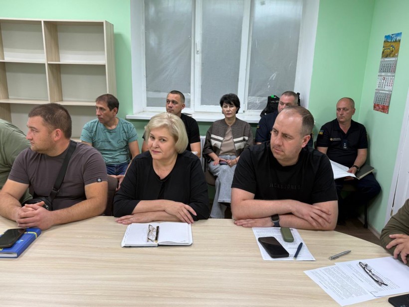 Учасники наради сидять за столом. На столі лежать відкриті блокноти та ручки. Всі уважно слухають.
