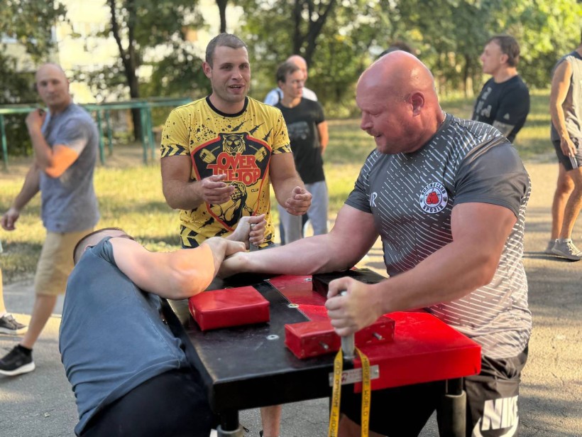 Двоє спортсменів змагаються на турнірі з  армрестлінгу. Поруч чоловік, який підтримує спортсменів.