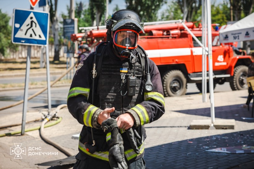 Позаду рятувальника стоїть пожежний автомобіль. Рятувальник у захисному одязі та маскою на обличчі, прямує уперед.