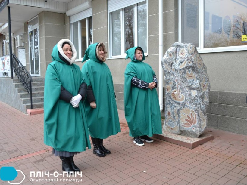 Троє жінок у зелених накидках стоять біля розписного каменю.