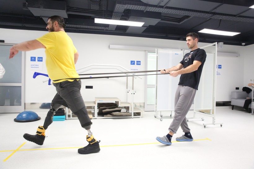 Двоє чоловіків перебувають у реабілітаційному центрі. Один з них одягнений у жовту сорочку і має протези ніг, енергійно ходить, а інший тягне за собою еластичні стрічки.