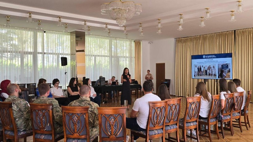 Світле приміщення. Стільці стоять кругом, на них сидять представники заходу. Молода жінка стоїть у центрі та розповідає, позаду неї екран з презентацією.