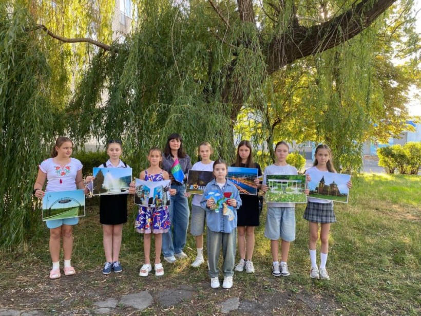Діти та педагог стоять під деревом з великими фотографіями визначних місць, спільна акція створює дух єдності та любові до рідного краю.