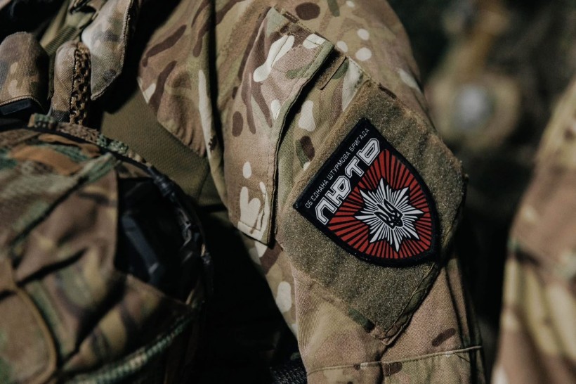 Збільшене зображення рукава військової форми в камуфляжі з помітною нашивкою з  текстом і емблемою у вигляді зірки, що передає відчуття авторитету.
