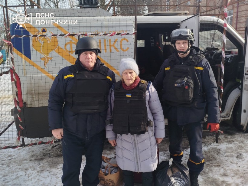 Рятувальники евакуаційної групи «Фенікс» та  літня жінка стоять біля броньованого автомобіля. Мешканка Дружківки з речами їде у безпечне місце.